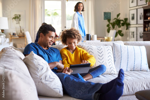 Close up of young father sitting on sofa in the living room with his pre-teen daughter using tablet computer, selective focus, mum in the background