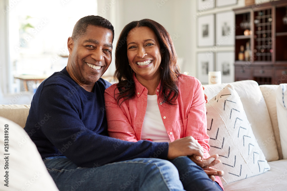 Middle aged mixed race couple sitting on the sofa in their living room looking to camera, front view, close up - obrazy, fototapety, plakaty 