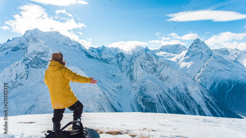 The guy stands on the ski slope in a yellow jacket with a snowboard. The guy is sitting on the ski slope in a yellow jacket with a snowboard. Mountain summit Dombai. National Park, Russia photo