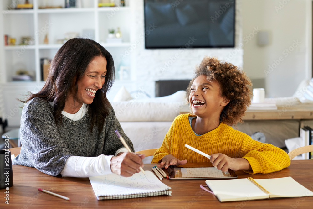 Middle aged mixed race woman sitting at a table in the dining room doing homework with her granddaughter, both laughing, close up