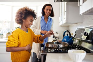 Pre-teen girl standing at hob in the kitchen using spatula and frying pan, preparing food with her mother, side view