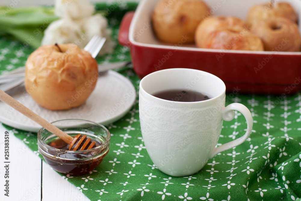 Baked apples in red, a white cup of tea or coffee, honey, a bunch of white tulips lie on a green tablecloth. Baked apples in red, a white cup of tea or coffee, honey, a bunch of white tulips lie on a green tablecloth.