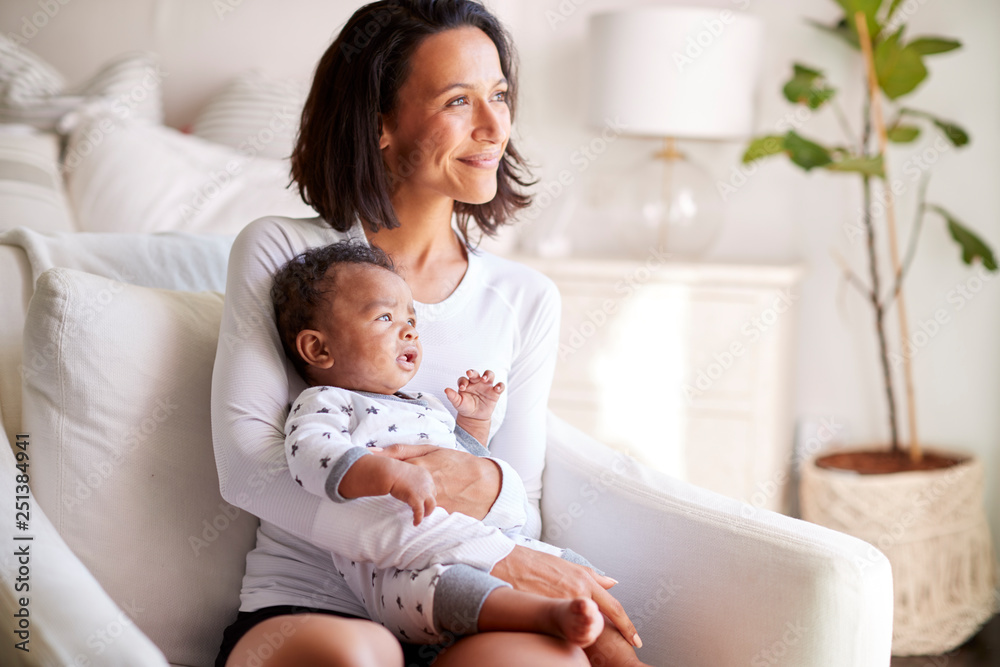 Young adult mother sitting in an armchair in her bedroom, holding her three month old baby son in her arms and looking away smiling, close up Young adult mother sitting in an armchair in her bedroom, holding her three month old baby son in her arms and looking away smiling, close up