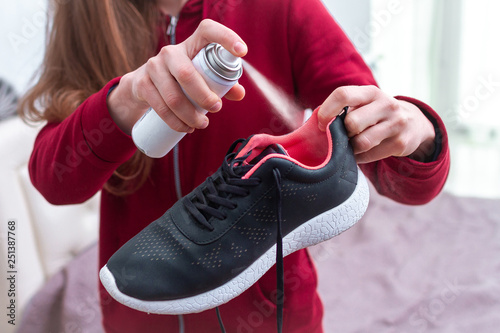 A young woman spraying deodorant on sweaty running shoes for eliminate unpleasant, bad smell. Shoe shine and care. Sport footwear needs in cleaning and odor removal.