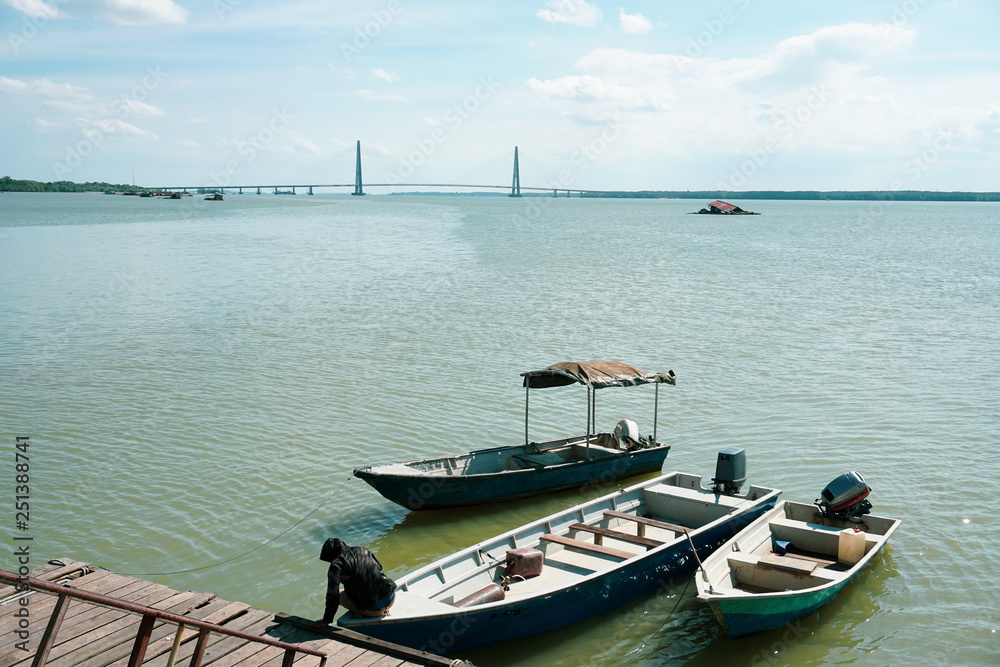 Fototapeta premium A man tie a rope to secure his boat to a wooden jetty with johor bridge in the background. view from a wooden jetty
