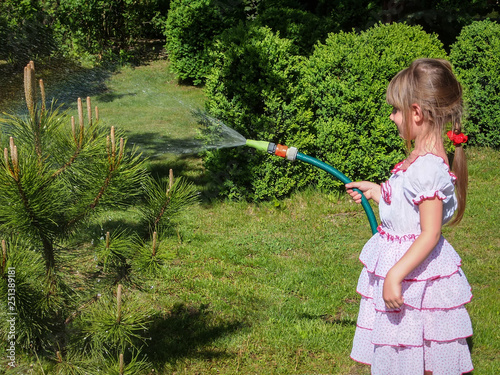 Pretty little girl 5 year old with long blond hair in lovelly white dress watering a small pine tree in the garden. Beautiful green background of spring garden. Ecology concept photo