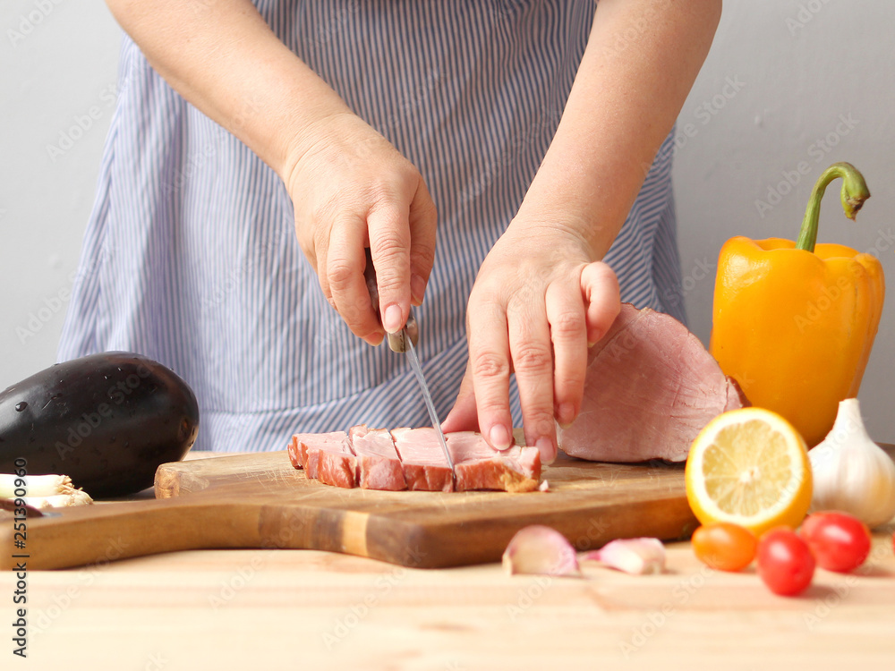 A woman cuts meat on a board, next to fresh vegetables lie: raw eggplant, carrots, lettuce, paprika, lemons, greens close-up on a light background
