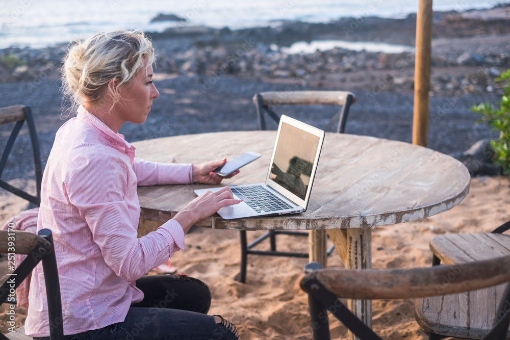 digital nomad concept with blonde free beautiful caucasian middle age woman working at the laptop using cellular phone too sitting on a wood table near the ocean - freelance free to work everywhere