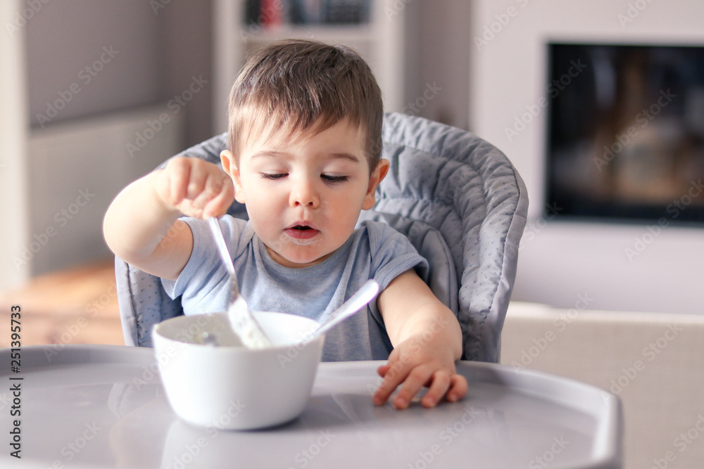 Cute little baby boy with funny smeared face concentrated on food eating with fork from white bowl at table in front of him sitting in high feeding chair. Attempt to eat by himself. Child nutrition Cute little baby boy with funny smeared face concentrated on food eating with fork from white bowl at table in front of him sitting in high feeding chair. Attempt to eat by himself. Child nutrition
