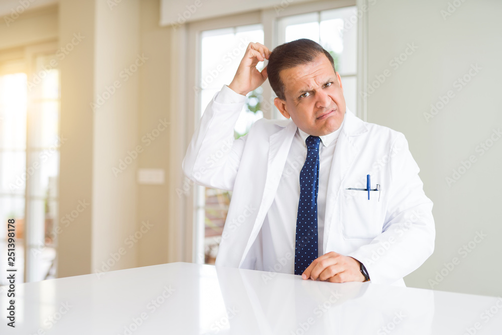 Middle age doctor man wearing medical coat at the clinic confuse and wonder about question. Uncertain with doubt, thinking with hand on head. Pensive concept. Middle age doctor man wearing medical coat at the clinic confuse and wonder about question. Uncertain with doubt, thinking with hand on head. Pensive concept.