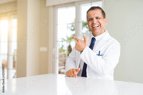 Middle age doctor man wearing medical coat at the clinic cheerful with a smile of face pointing with hand and finger up to the side with happy and natural expression on face