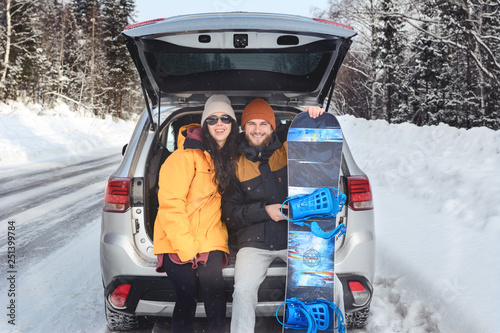 Smiling couple is sitting in the back trunk of suv car on roadside of winter road. Family trip to ski resort. Winter holidays adventure.  Man holding his snowboard. travelling lifestyle concept.