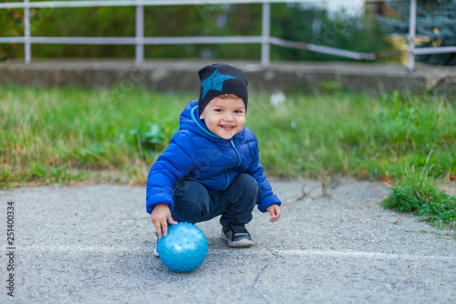 a boy in a blue jacket is sitting in the street with a ball. smile and laugh.