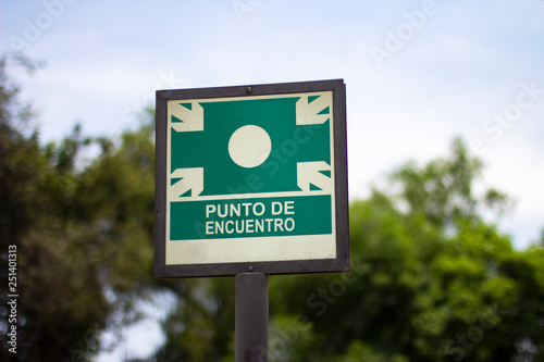 Street sign of "punto de encuentro" meeting point against green trees in spanish. Emergency evacuation assembly point signboard.