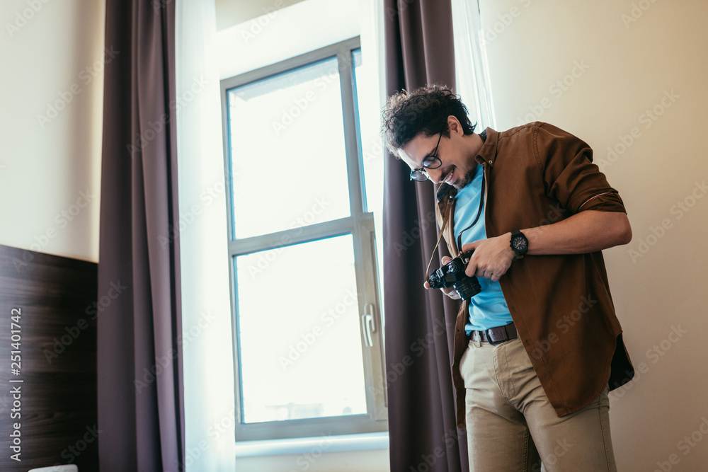 smiling man in eyeglasses looking at photo camera near window in hotel room