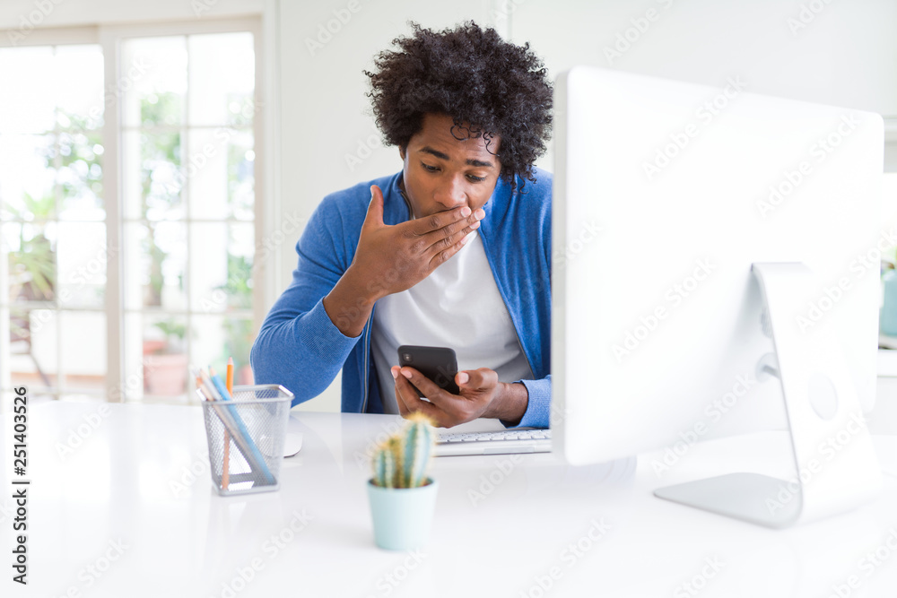 African American man working using smartphone and computer cover mouth with hand shocked with shame for mistake, expression of fear, scared in silence, secret concept African American man working using smartphone and computer cover mouth with hand shocked with shame for mistake, expression of fear, scared in silence, secret concept