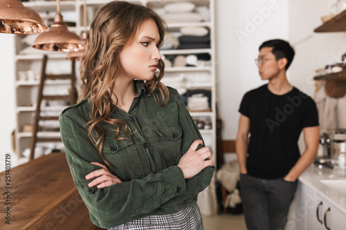 Upset woman with wavy hair holding hands together sadly looking aside with asian man on background. Young international couple quarreling while spending time on kitchen at home