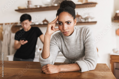 Angry african american woman holding hand near head sadly looking aside with asian man using cellphonr on background. Young international couple in quarrel spending time on kitchen photo