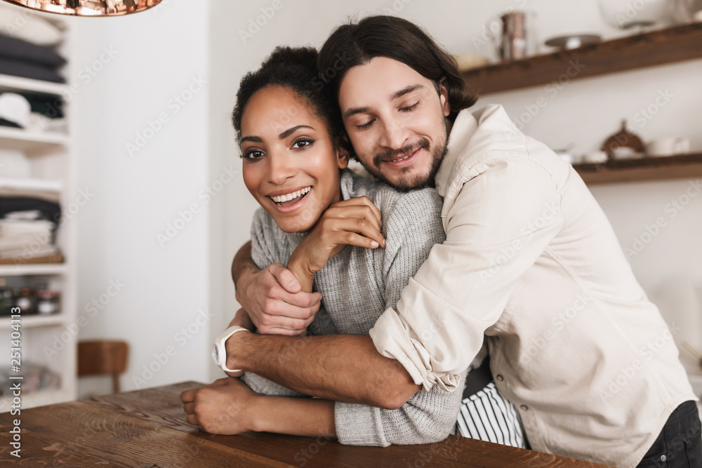 Handsome man dreamily embracing beautiful african american woman. Young international couple happily spending time together in cozy kitchen at home - obrazy, fototapety, plakaty 