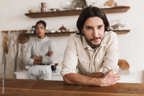 Attractive man with beard leaning on table while thoughtfully looking in camera with african american woman on background. Young international couple in quarrel spending time in kitchen at home