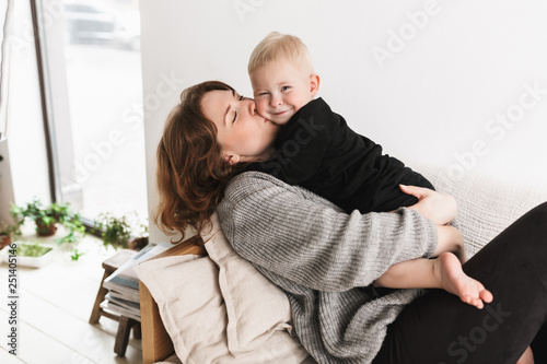 Young pretty woman lying on sofa dreamily kissing her little handsome son in cheek that happily looking in camera. Mom with baby boy joyfully spending time in living room at home