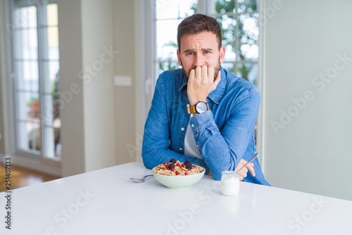 Handsome man eating cereals for breakfast at home looking stressed and nervous with hands on mouth biting nails. Anxiety problem.