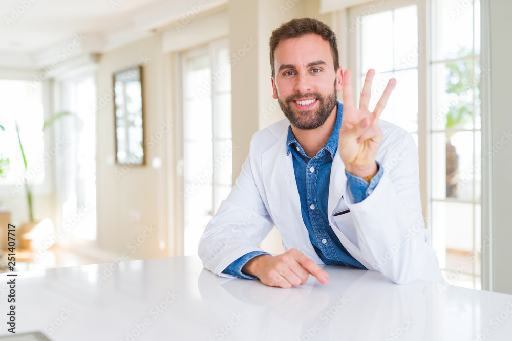Handsome doctor man wearing medical coat at the clinic showing and pointing up with fingers number three while smiling confident and happy.