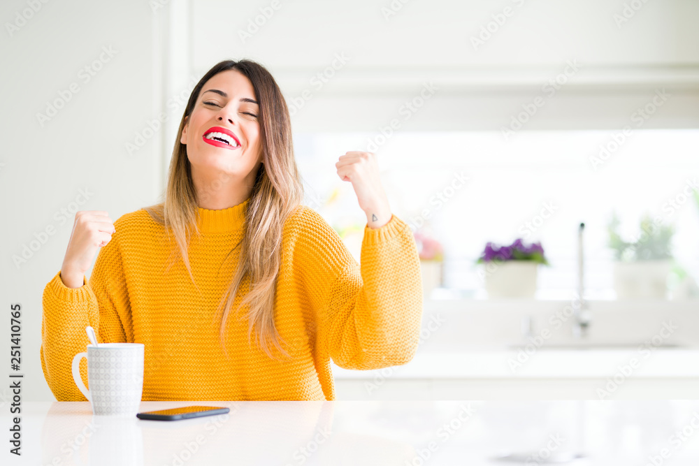 Young beautiful woman drinking a cup of coffee at home very happy and excited doing winner gesture with arms raised, smiling and screaming for success. Celebration concept. Young beautiful woman drinking a cup of coffee at home very happy and excited doing winner gesture with arms raised, smiling and screaming for success. Celebration concept.