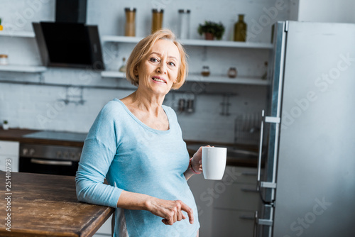 smiling senior woman with cup of coffee looking at camera in kitchen
