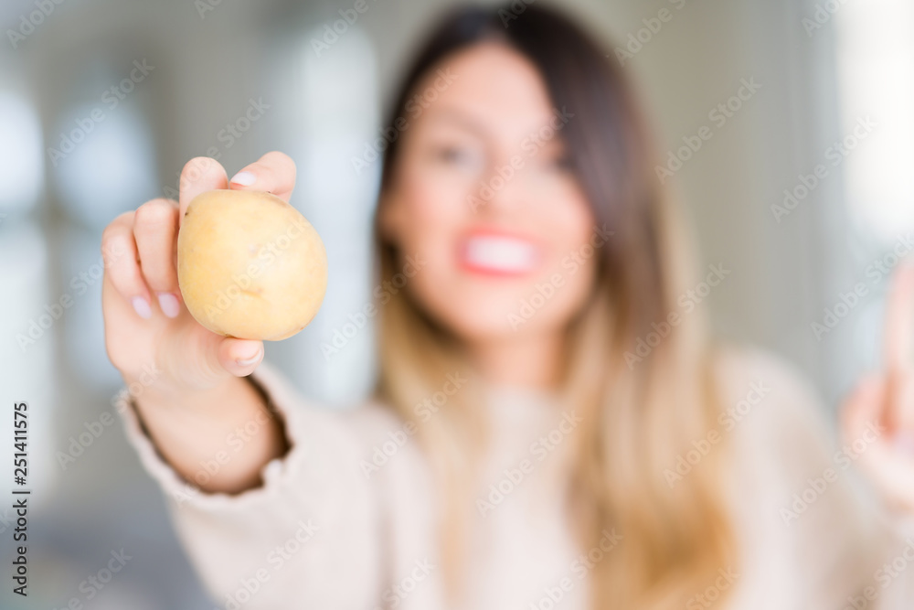 Young beautiful woman holding fresh potato at home very happy pointing with hand and finger to the side Young beautiful woman holding fresh potato at home very happy pointing with hand and finger to the side