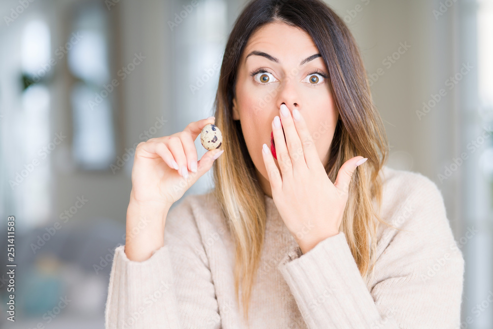 Young beautiful woman holding fresh quail egg at home cover mouth with hand shocked with shame for mistake, expression of fear, scared in silence, secret concept Young beautiful woman holding fresh quail egg at home cover mouth with hand shocked with shame for mistake, expression of fear, scared in silence, secret concept