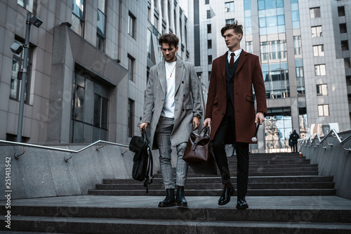 Two fashion men models posing on city street. Wearing in classic and casual closes. Suit, coat, shirt, sweater, boots and leather bag. Men model test near business center