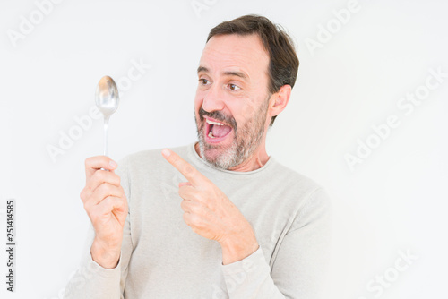 Senior man holding silver spoon over isolated background very happy pointing with hand and finger