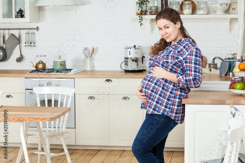 Pregnant woman holding her tummy with hands, standing close to countertop in kitchen, copy space