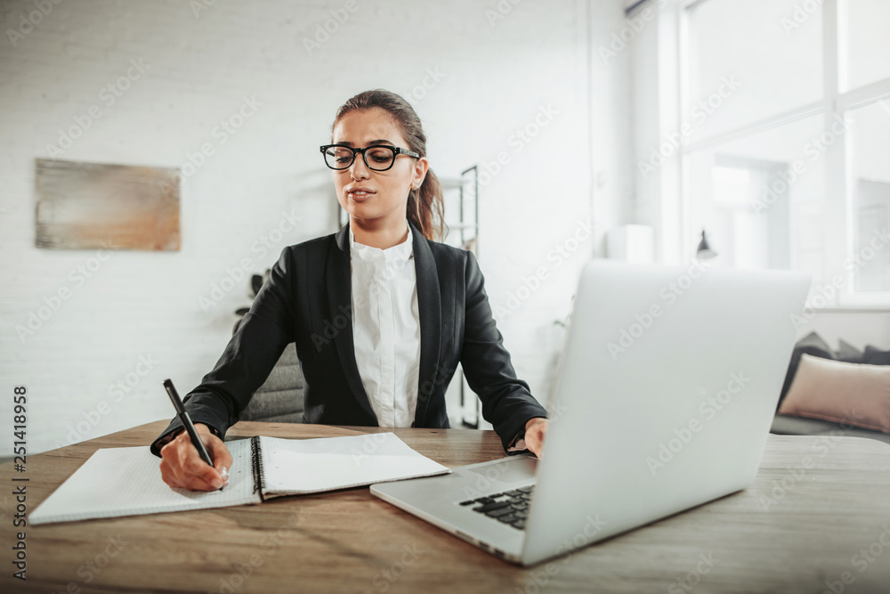 Smart and serious girl sits at table and works. She writes down information in notebook. The other hand in on keybpard. Girl wears glasses