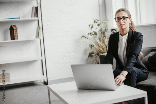 Positive and nice girl sits on sofa and looks on camera. She holds her hands on keyboard. Girl is in white office with plants and books photo