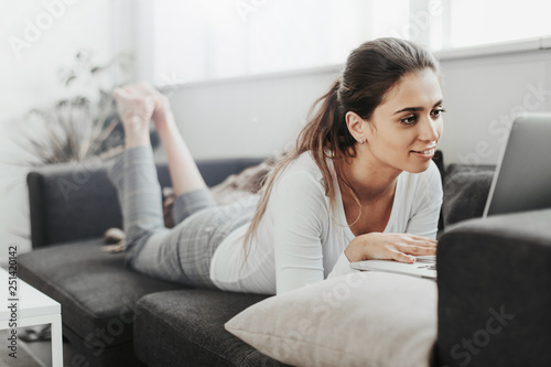 A picture of nice and calm girl lying on stomacj on sofa. She works. Woman looks at laptop. She in concentrated. Girl is in white room with dark decoration