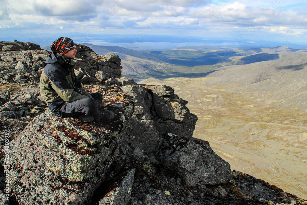 Caucasian white male tourist in sportswear, buff and glasses sits on a rock against the backdrop of mountains Caucasian white male tourist in sportswear, buff and glasses sits on a rock against the backdrop of mountains
