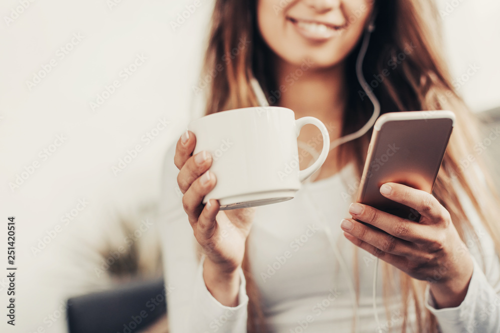 Cut view of a girl holds white cup and phone. She looks at it and smiles. Woman is listening to music. She is very attractive Cut view of a girl holds white cup and phone. She looks at it and smiles. Woman is listening to music. She is very attractive