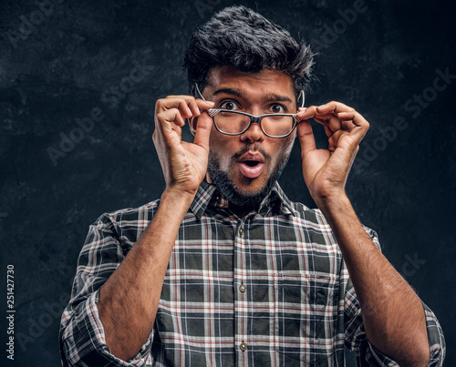 Surprised Indian guy lowers his glasses and looks at the camera. Studio photo against a dark textured wall