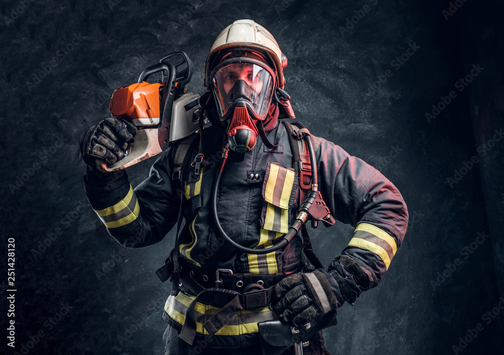 Naklejka premium A brave firefighter wearing full protective equipment posing with a chainsaw on his shoulder. Studio photo against a dark textured wall