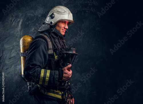 Brutal firefighter in uniform and safety helmet holding an oxygen mask and looking sideways with a confident look.