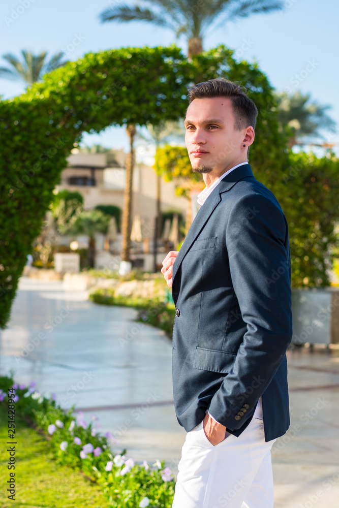 Young handsome man in man in a white shirt, jacket and white trousers at the palm trees background on a beach. Young handsome man in man in a white shirt, jacket and white trousers at the palm trees background on a beach.