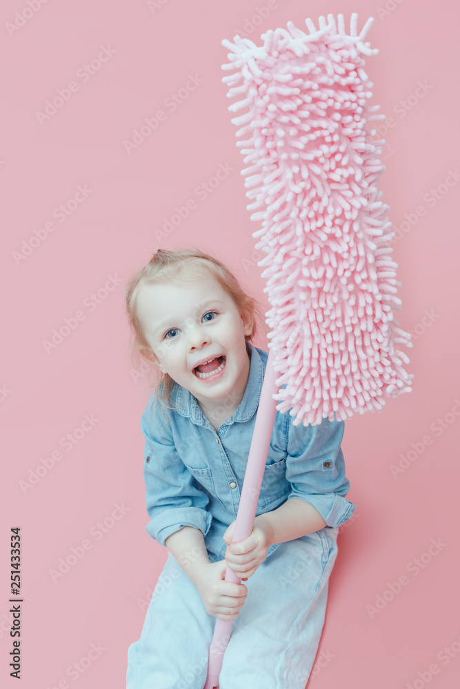 A small child in denim clothes sits on a pink background and holds a pink MOP.