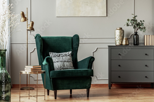 Emerald green armchair with pillow next to grey wooden commode in dark living room interior