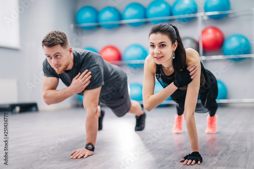 Beautiful woman and strong male doing push-ups on one hand with a background of fitness balls. Young couple is working out at gym
