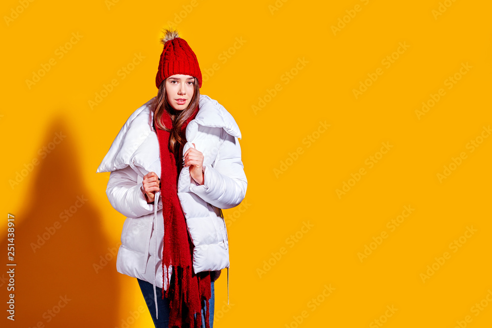 Stylish young woman in a white down coat and knite red hat on yellow background in studio. Stylish young woman in a white down coat and knite red hat on yellow background in studio.
