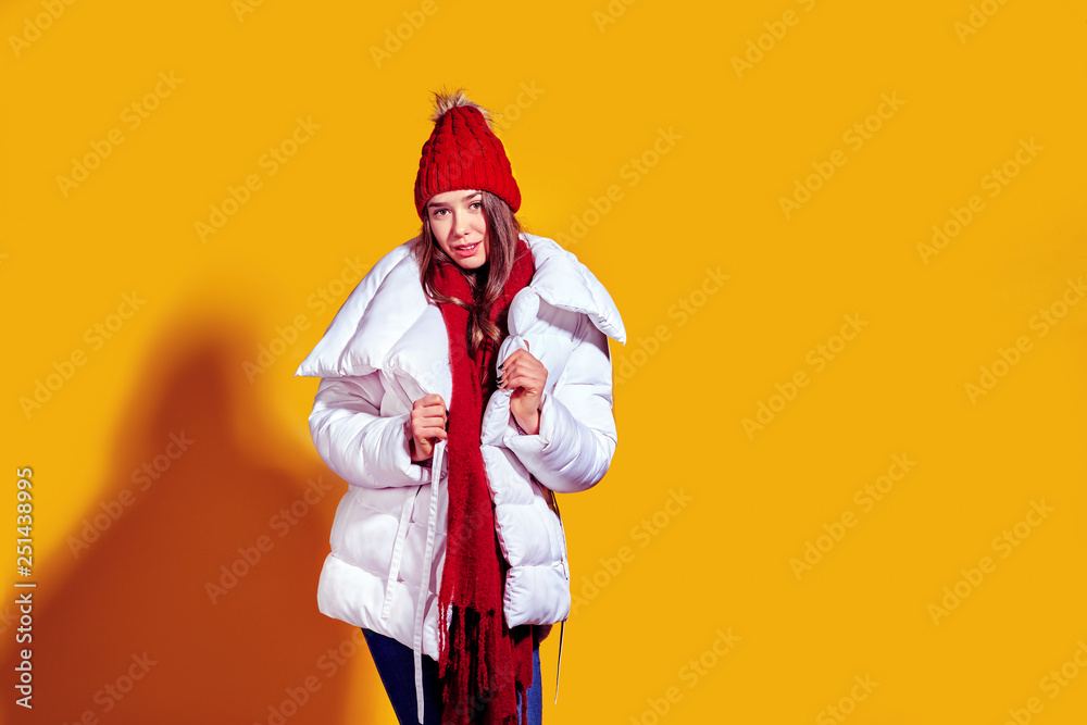 Stylish young woman in a white down coat and knite red hat on yellow background in studio.