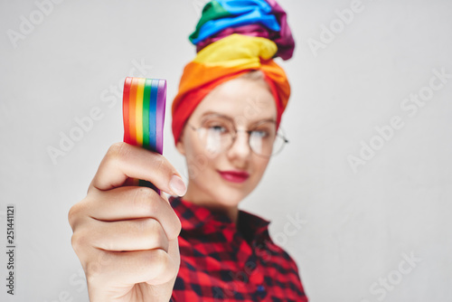 A beautiful young woman with glasses and full lips in a turban and a red shirt holds a rainbow in her hand against a white wall. LGBTQ Lesbians, gays, bisexuals, transgender, queer. Homosexual man