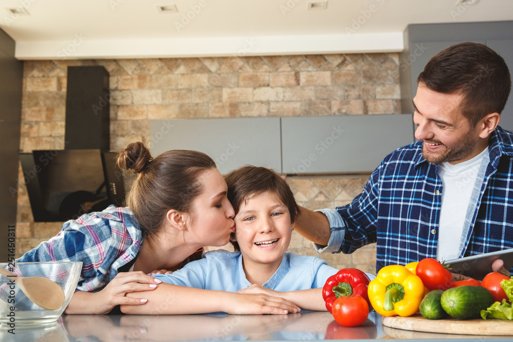 Family at home standing in kitchen together mother kissing son while father touching his head praising satisfied Family at home standing in kitchen together mother kissing son while father touching his head praising satisfied